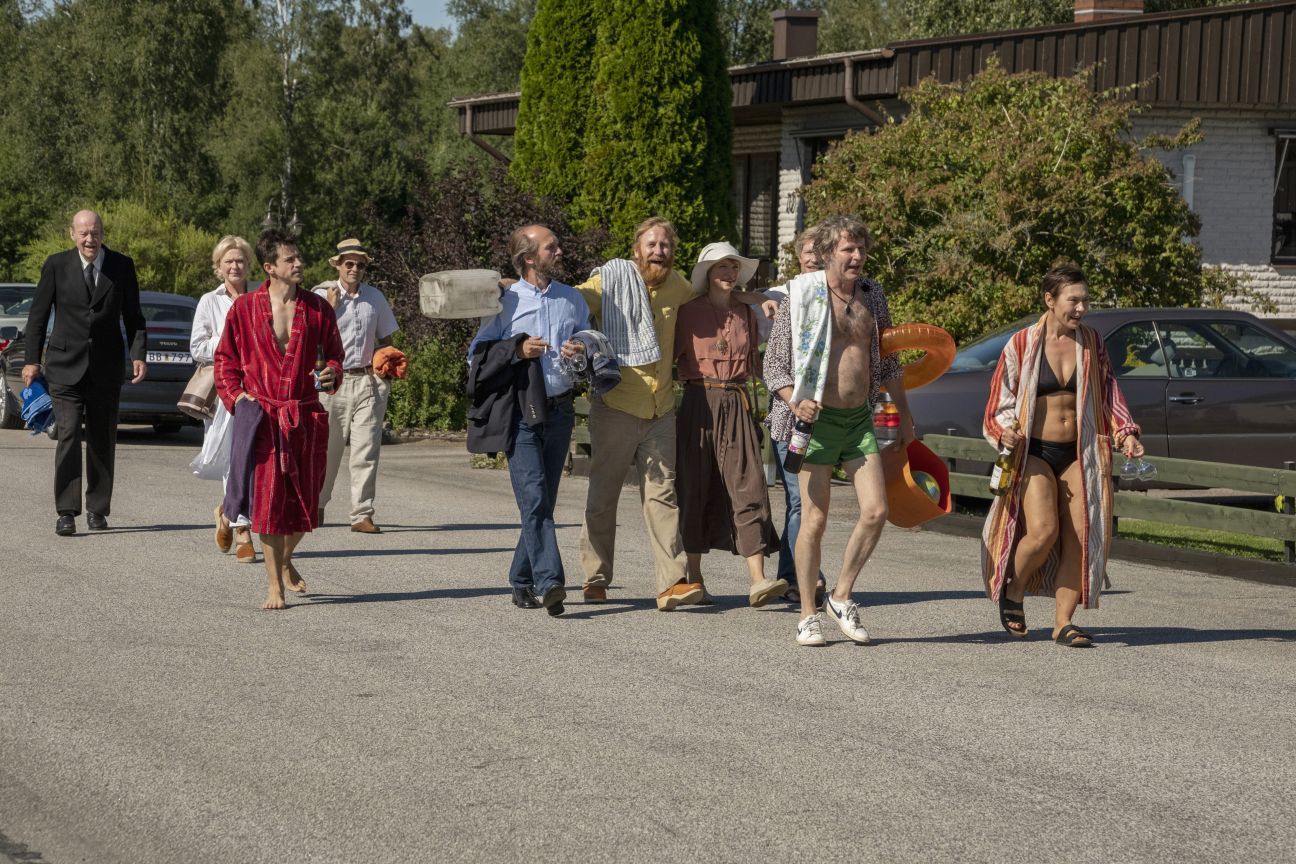 Bildbeschreibung: Eine Gruppe Menschen, teilweise in Badekleidung, geht eine Straße hinunter. Standbild aus dem Film "Tilsammans 99".