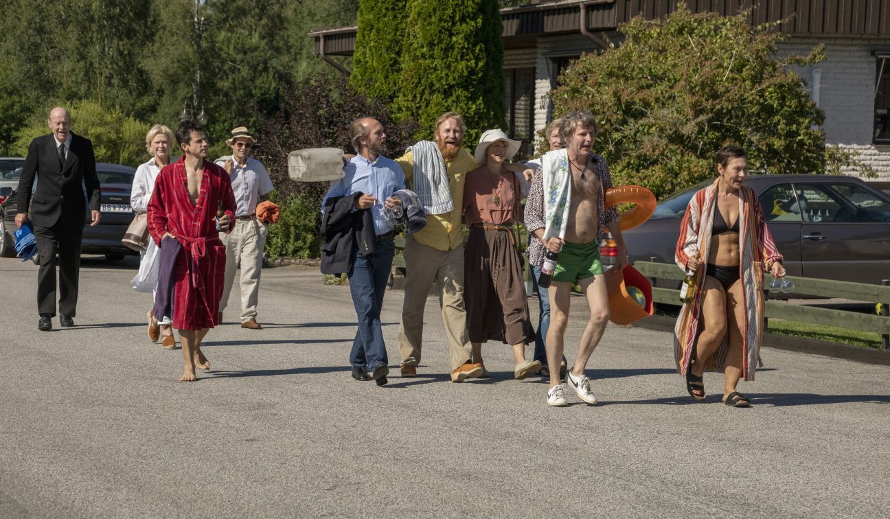 Bildbeschreibung: Eine Gruppe Menschen, teilweise in Badekleidung, geht eine Straße hinunter. Standbild aus dem Film "Tilsammans 99".