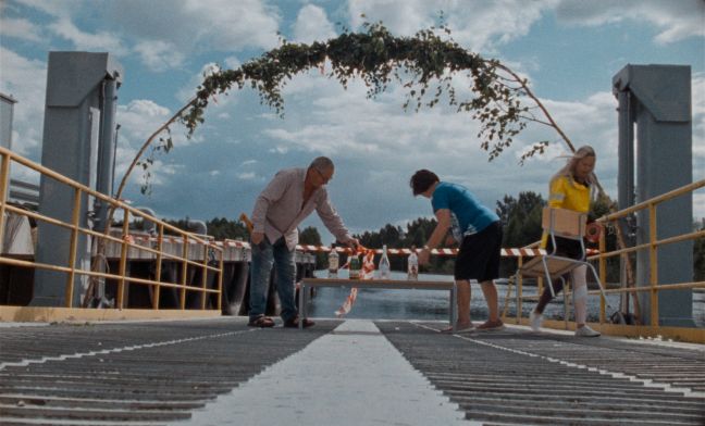 Drei Menschen stehen vor einer abgesperrten Bootsrampe mit einem Stuhl und einem Tisch, worauf verschiedene Spirituosenflaschen stehen