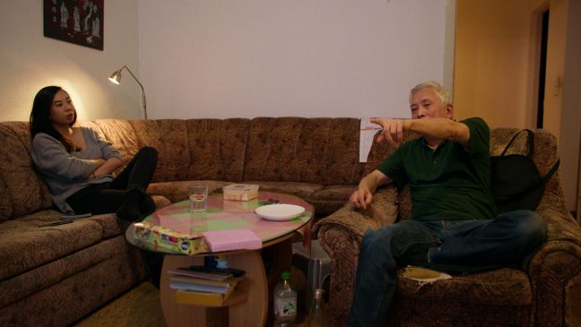 Anna sitzt auf dem Sofa, gegenüber von Vater Hung auf dem Sessel, der in die Ferne zeigt