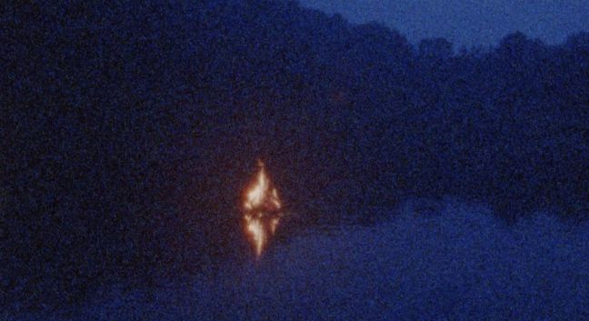 Nachtaufnahme von einem Wald, davor brennt ein offenes Feuer, rauschendes Bild