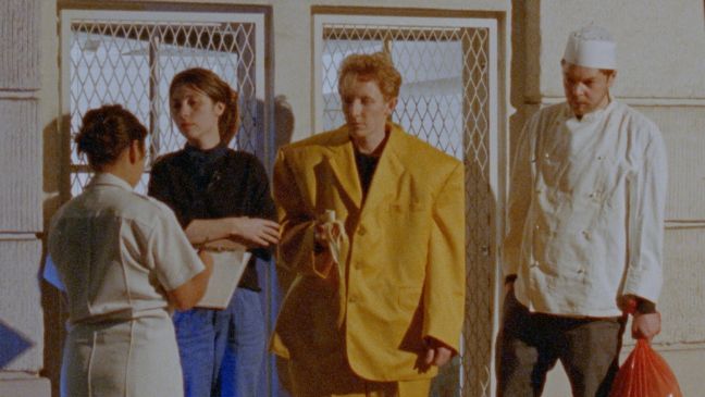 Ein Schauspieler in breitem gelben Anzug und einer in Küchenuniform stehen neben zwei Frauen der Crew