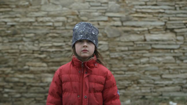 Roosi mit acht Jahren im Wind mit Mütze und roter Jacke vor einer Steinwand