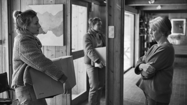 Die Tochter steht mit einem Karton in der Hand in der Haustür, die Mutter schaut sie mit verschränkten Armen an