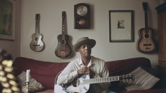 Eric Bibb sitzt auf einer roten Couch mit Gitarre auf dem Schoß, Gitarren hängen hinter ihm an der Wand