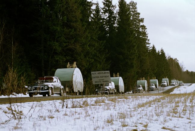 Eine Kolonne von gleichen Volvo PV mit Wohnwagen fährt eine Straße neben einem schneebedecktem entlang