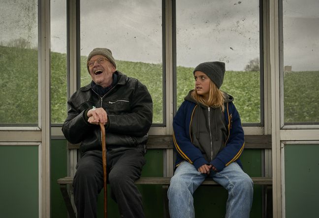 Honey entsetzt, Marcel lachend, sitzen auf der Bank in einer Bushaltestelle