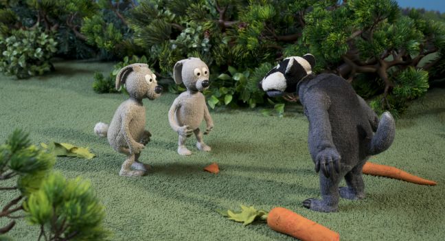 Der Dachs steht zwischen zwei Karotten und baut sich vor den zwei Hasen auf