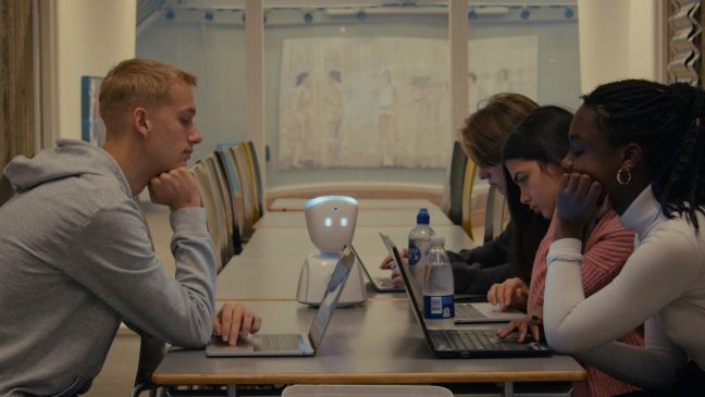 vier Jugendliche mit Laptops sitzen an einem Tisch, darauf steht ein kleiner Roboter