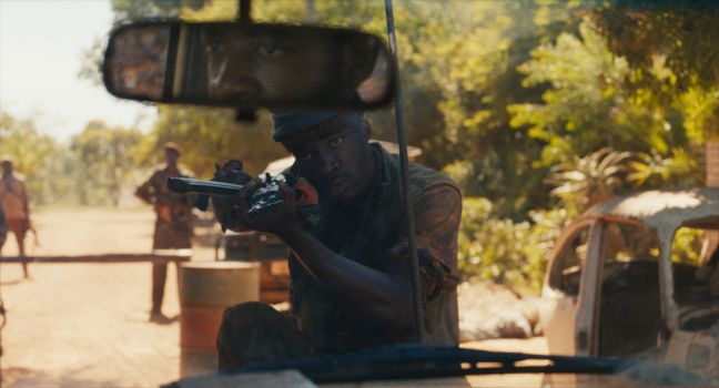 Bewaffneter Mann zielt in die Ferne, im Rückspiegel des Autos das Gesicht des Fahrers