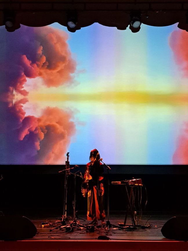 Veera Neva auf der Bühne mit Instrumenten, hinter ihr die Leinwand mit bunten Wolken