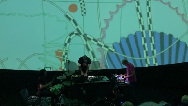 Saroos spielt vor der Leinwand auf ihren Instrumenten: Schlagzeug, Keyboard und Synthesizer