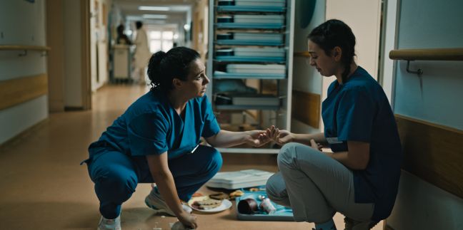 Melina hockt weinend am Boden, daneben heruntergefallene Tabletts mit Essen, Teresa vor ihr, tröstend