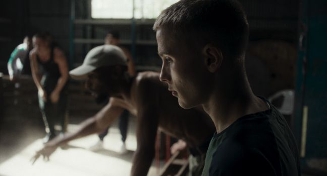 Robin schaut auf den Boden, im Hintergrund gestikulieren Männer