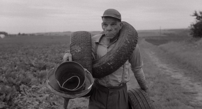 Karl-Göran läuft auf dem Feldweg, drei Autoreifen über seinen Körper gelegt mit verschiedenen Metallteilen in der Hand