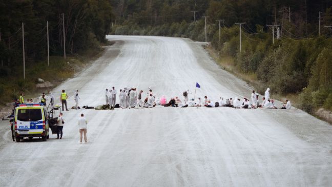 Auf einer breiten Sandstraße bilden Demonstranten eine Blockade, links ein Fahrzeug der Dialogpolizei