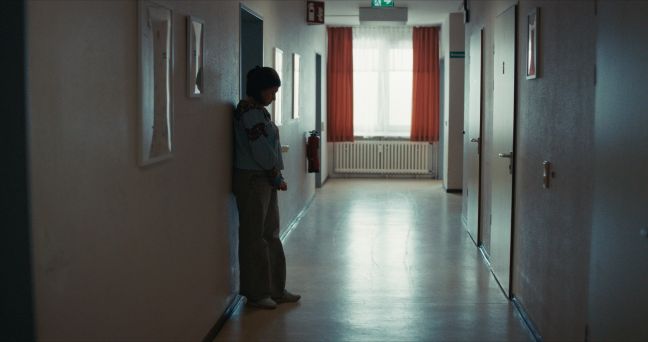 Luisa steht an die Wand gelehnt, die Hände verschränkt, Blick auf den Boden