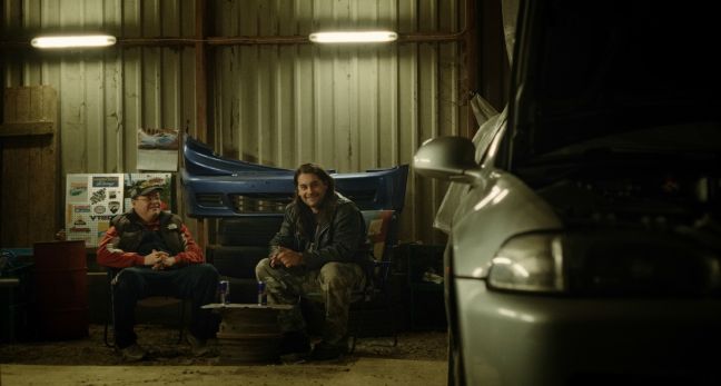Martin und Casper trinken Energy Drinks in der Garage; im Vordergund ein Auto
