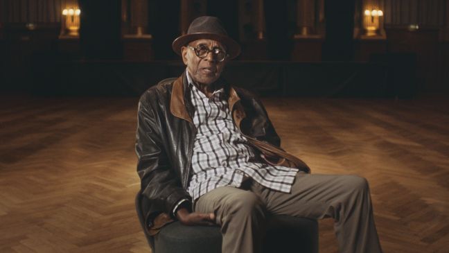 Albert Heath sitzt auf einem Stuhl in einem dunkel beleuchteten 
historischen Saal