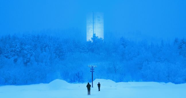 Zwei Menschen stehen im Schnee vor einem Wegweiser in der Weite eines Waldes, blaustichiges Bild