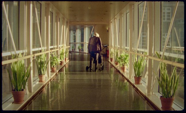 Aufnahme von hinten, Protagonistin geht mit den Rollator  durch den voll Licht-durchfluteten Flur