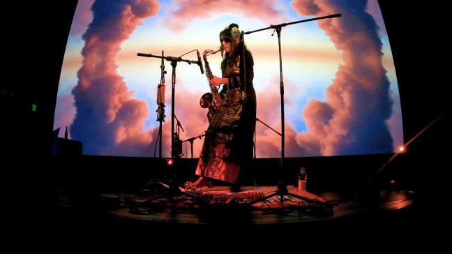 Veera Neva spielt Saxophon auf der Bühne, auf der Leinwand hinter ihr sind rötliche Wolken