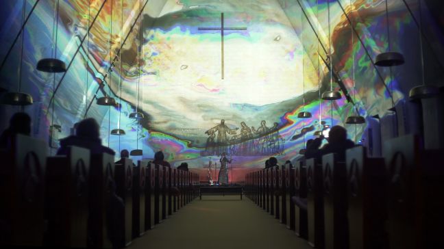 Veera Neva in der Ferne mit Mikrofon, ein Saal der durch Prisma-Linien und religiösen Symbolen gefüllt ist