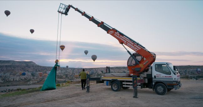 Eine Frau mit langem grünen Kleid wird mithilfe eines Krans auf einer Schaukel in die Luft gehoben, im Hintergrund zerklüftete Hügel und Heißluftballons