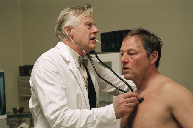 Grant wird mit freiem Oberkörper von Dr. Benjaminsen untersucht, während dieser raucht