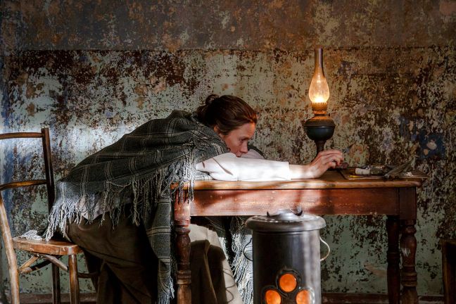 Eine Frau sitzt auf der Kante des Stuhls, der Oberkörper über den Tisch, auf dem eine Öllampe steht