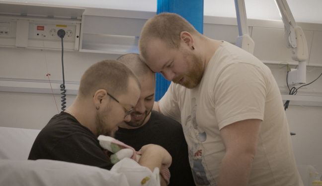 Kristopher, David und Sindre halten das Baby im Krankenhaus und schauen es liebevoll an
