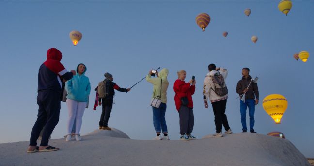 Auf einem sandigen Hügel stehen Menschen und fotografieren den Himmel voller Heißluftballons