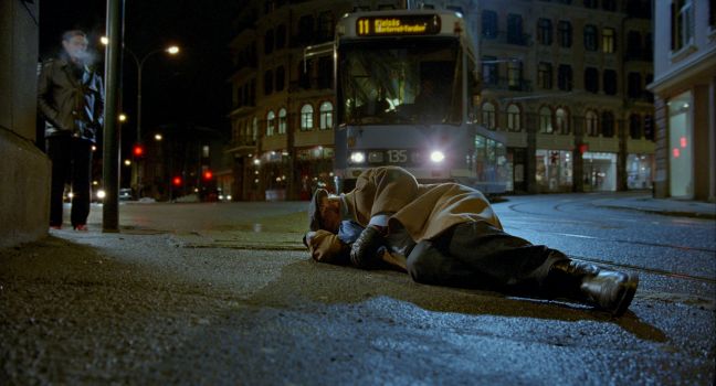 Ein alter Mann liegt auf der Straße neben den Straßenbahngleisen, die Straßenbahn fährt dahinter