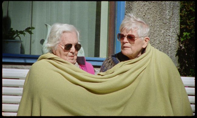 Der Protagonist und die Protagonistin sitzen draußen in der Sonne zugedeckt mit einer Decke und tragen Sonnenbrille