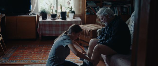 Miia kniend, zieht dem Großvater die Hose an