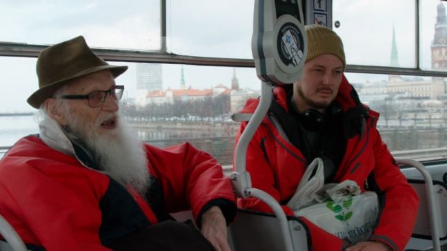 Margers fährt in roter Jacke mit dem Regisseur im Bus