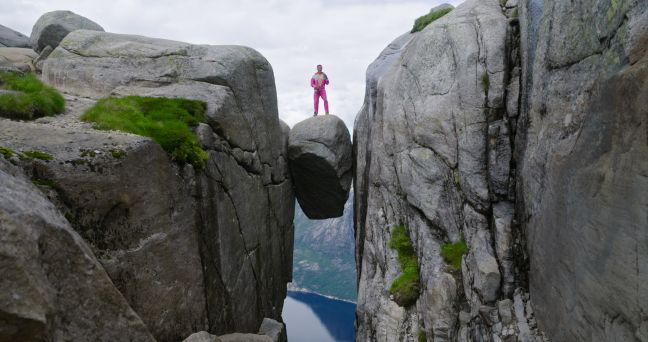 Ein Mann in magentafarbigen Anzug posiert auf einem Fels, der in einer Spalte eingeklemmt ist über dem Abgrund