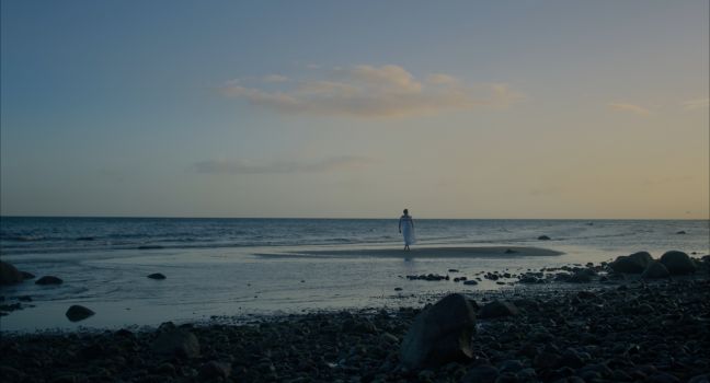 Paul in weißem Sommerkleid auf einer Sandbank im Wasser, im Anschnitt die steinige Küste