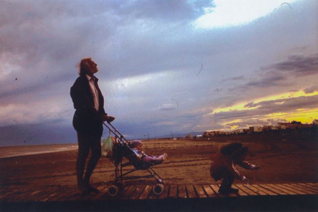 Am Meer, Vater schiebt Wagen mit Baby und die andere Tochter bückt sich