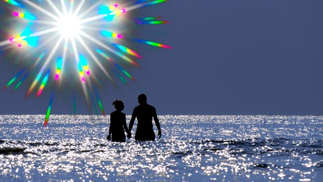 Ein Mann und eine Frau im Wasser im Gegenlicht, die Sonne spaltet sich in Prismafarben aus