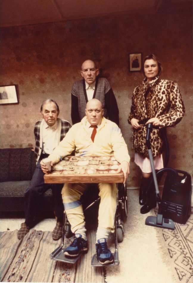 Moe sitzt neben und Far steht hinter Konrad, der im Rollstuhl einen Sortierkasten mit Eiern auf dem Schoß hat, Cylindia mit einem Staubsauger daneben, alle schauen in die Kamera