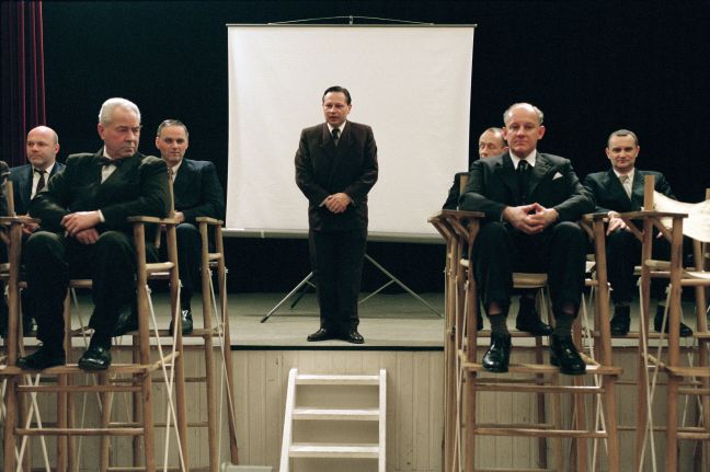 Malmberg steht mittig vor einer Leinwand, umgeben von Männern in Anzügen, die in Hochstühlen sitzen