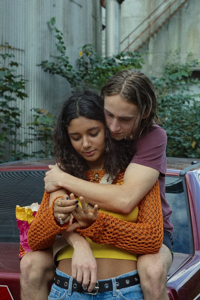 Alex umarmt Ines von hinten sitzend auf der Motorhaube