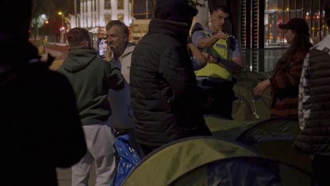 Menschen die sich mit dem Handy filmen, ein Polizist, dazwischen Zelte