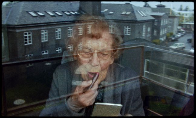 Protagonistin schminkt sich vor dem Fenster, ein Gebäude ist durch das Fenster sichtbar