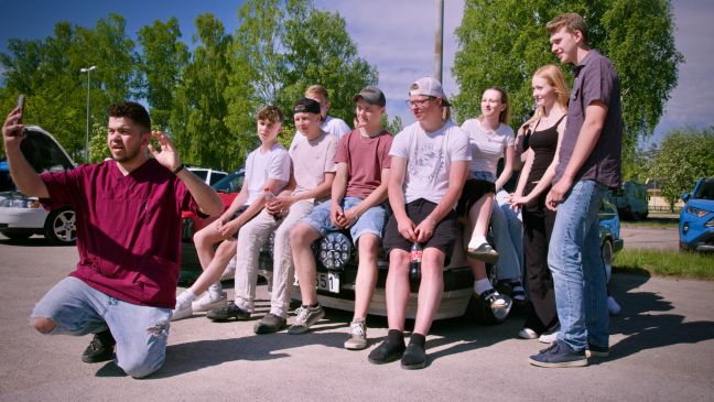 Majd nimmt mit seinem Smartphone ein Video auf, kniend vor einer Gruppe Teenager, die auf einem Auto sitzen