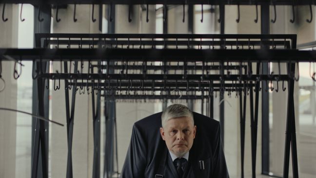 Der Mann steht unter einem Anzug, der an der Garderobe hängt, sodass sein Körperbau deformiert wirkt