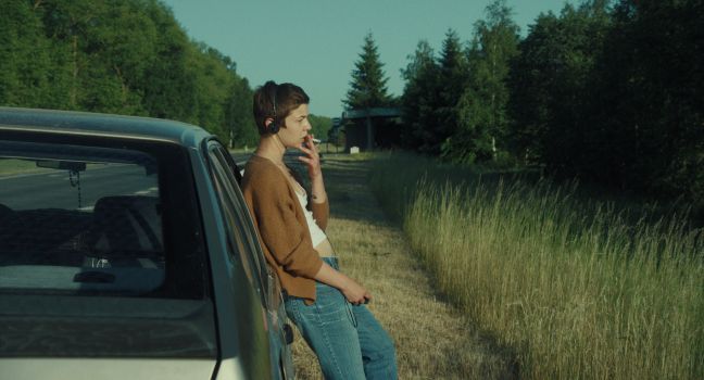 Ada lehnt an dem Auto, raucht und hat Kopfhörer auf