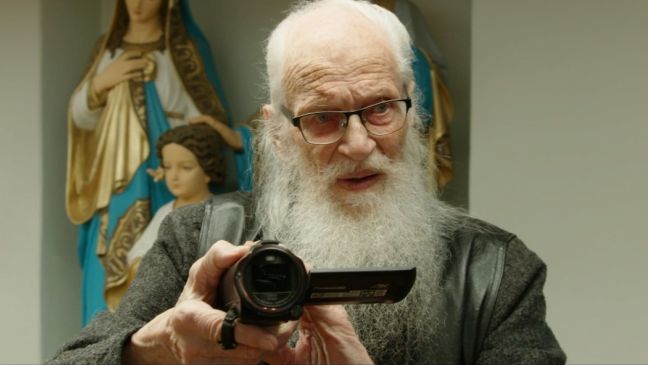 Margers frontal, filmt mit einer Videokamera, im Hintergrund Statuen in goldenem Gewand