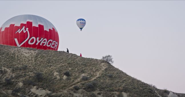 Über einem Hügel schwebt ein ferner Heißluftballon, ein naher kommt hinter dem Hügel hervor
